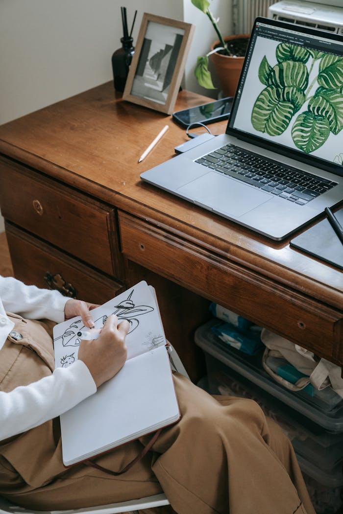 A person sketching in a notebook at a wooden desk with a laptop displaying digital art.